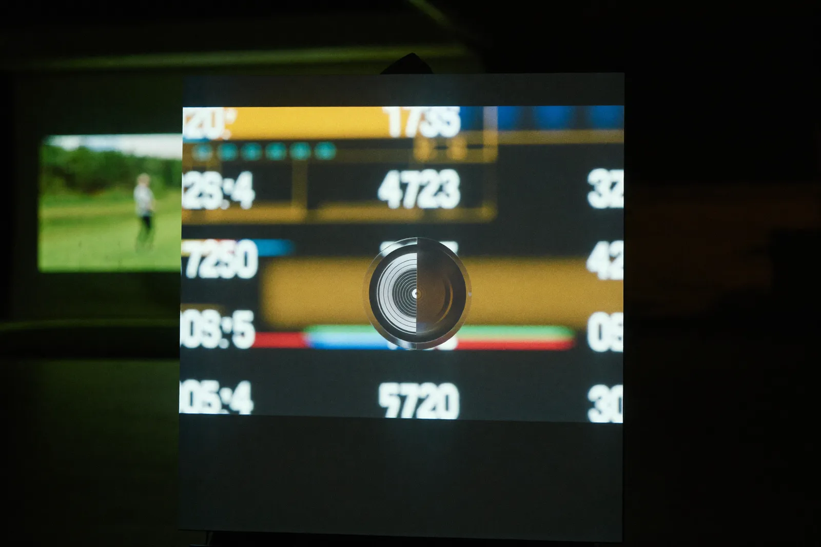 A launch monitor glowing on the floor of a dark garage, a golfer silhouetted beyond