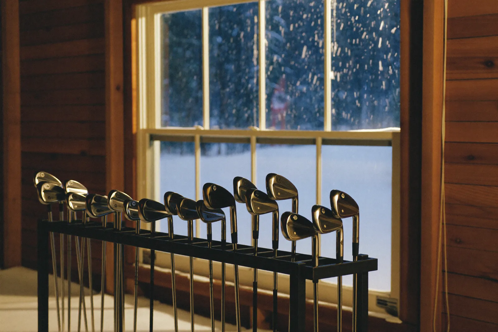A rack of golf irons in a tidy garage, snow falling outside the window behind them