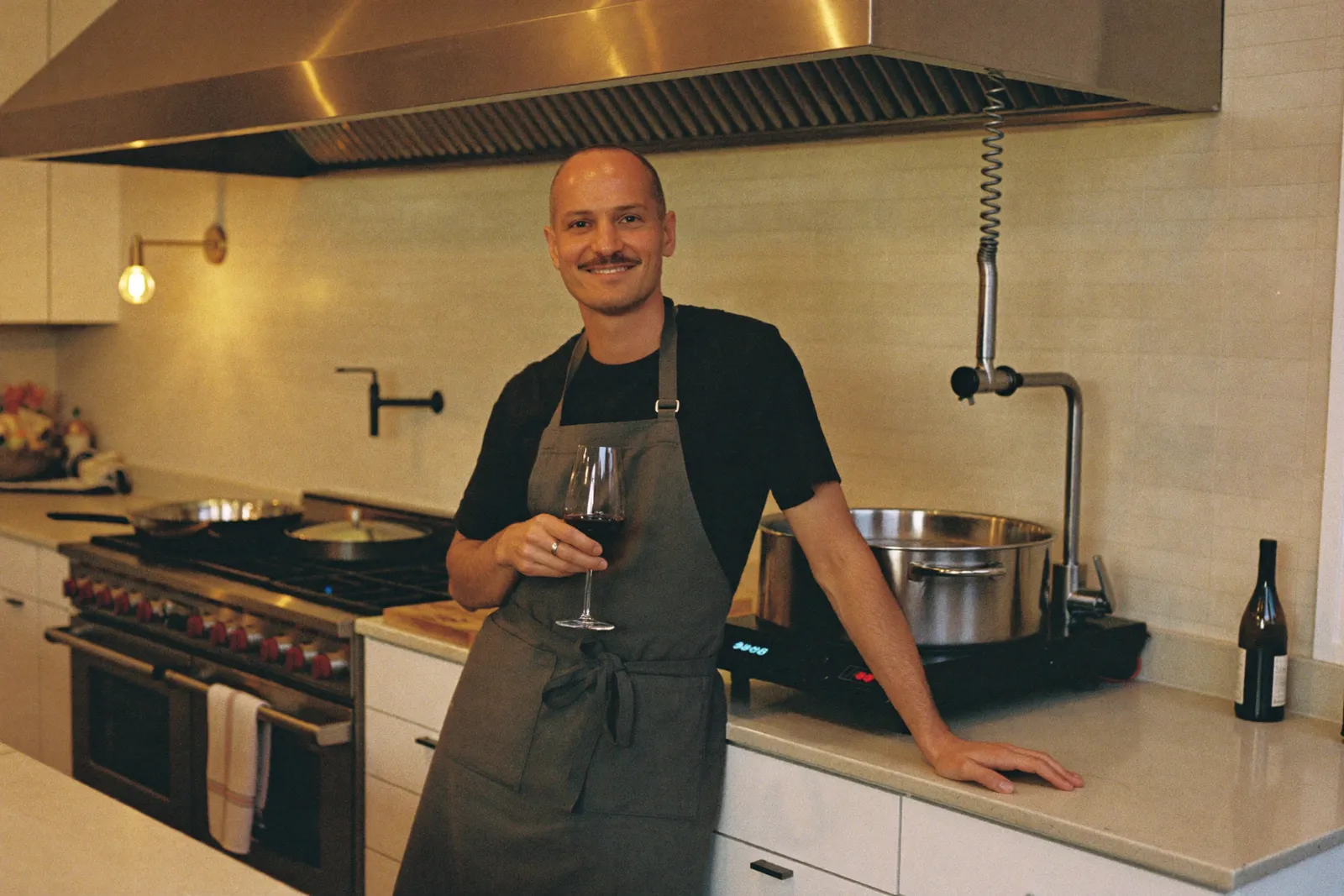 A relaxed man in an apron leaning against the counter with a glass of red wine while a sous vide bath works behind him