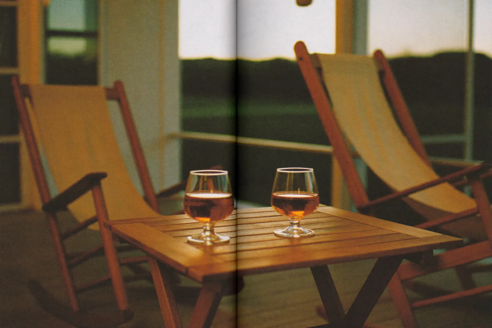 Two tulip whiskey glasses on a porch table at dusk, two rocking chairs angled toward each other