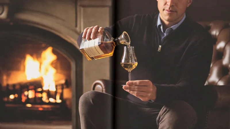 A man pouring whiskey from a decanter into a tulip glass beside a stone fireplace