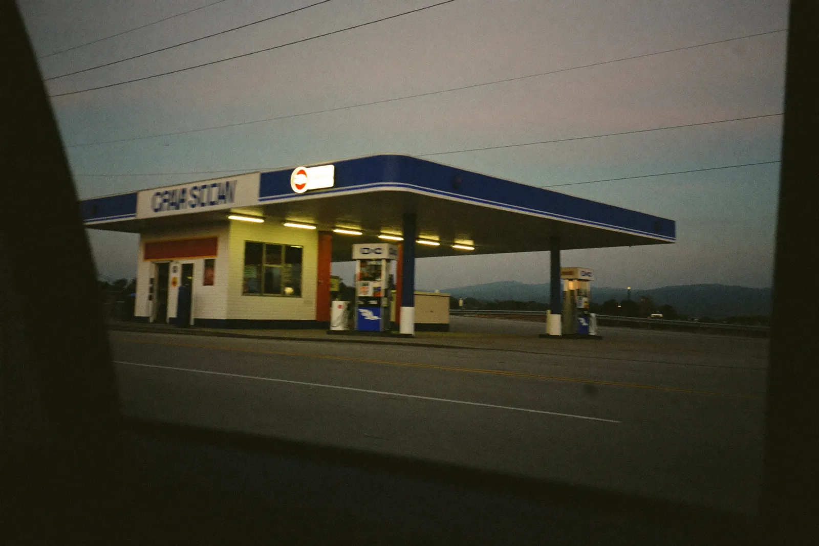 An old roadside gas station at dusk, seen through the window of a passing car
