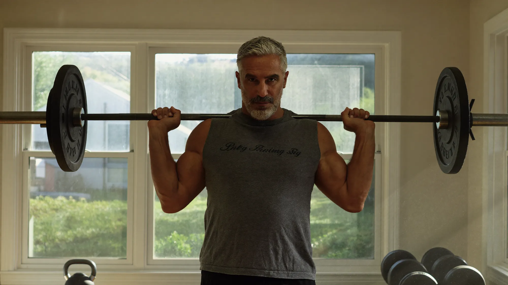 A silver-haired man setting up a barbell deadlift in a clean home gym, morning light through the window