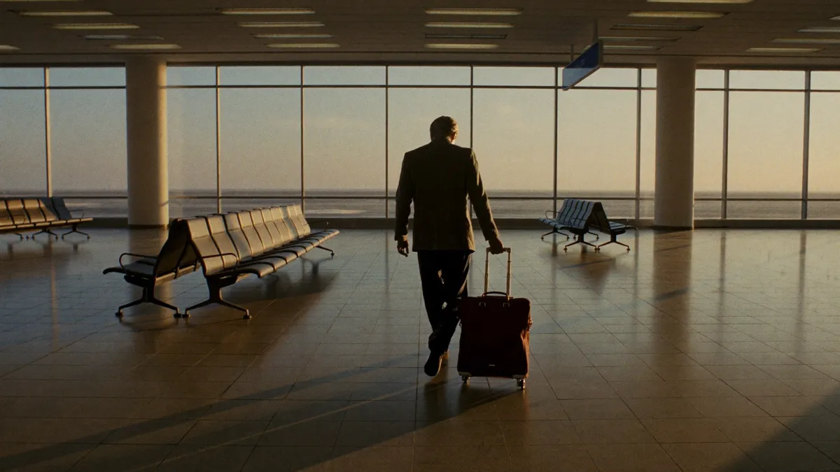 A silver-haired man pulling one small roller bag through an empty airport concourse at dawn