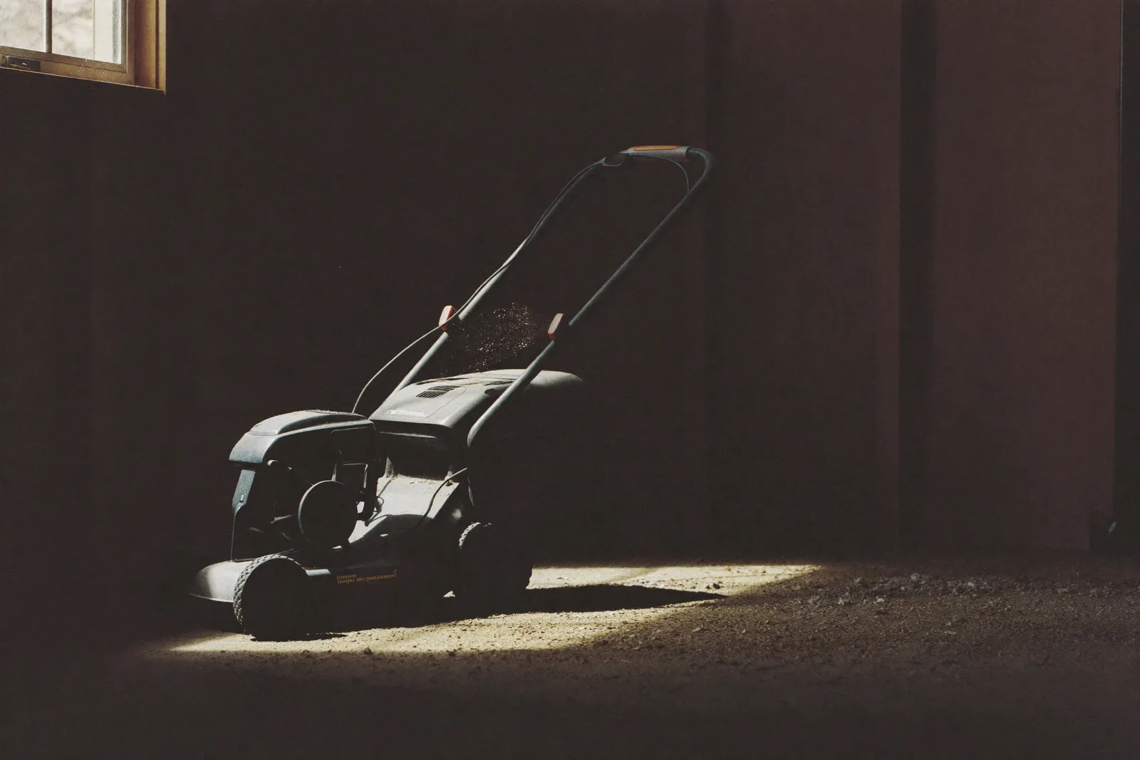 A dusty push lawnmower in the dark corner of a garage, lit by a single shaft of window light