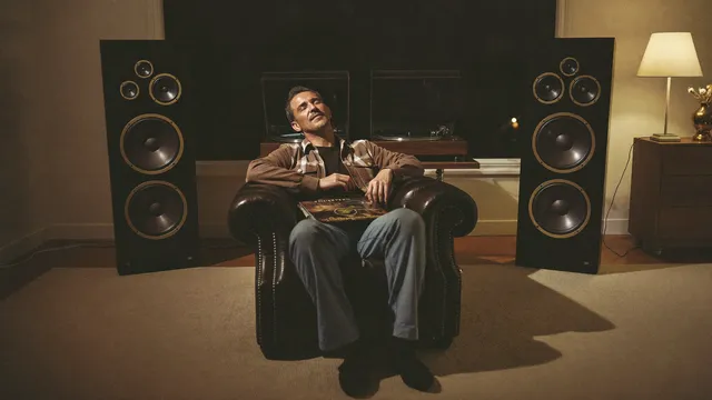 A man in a leather armchair between two floor-standing speakers in a warm study at night, eyes closed