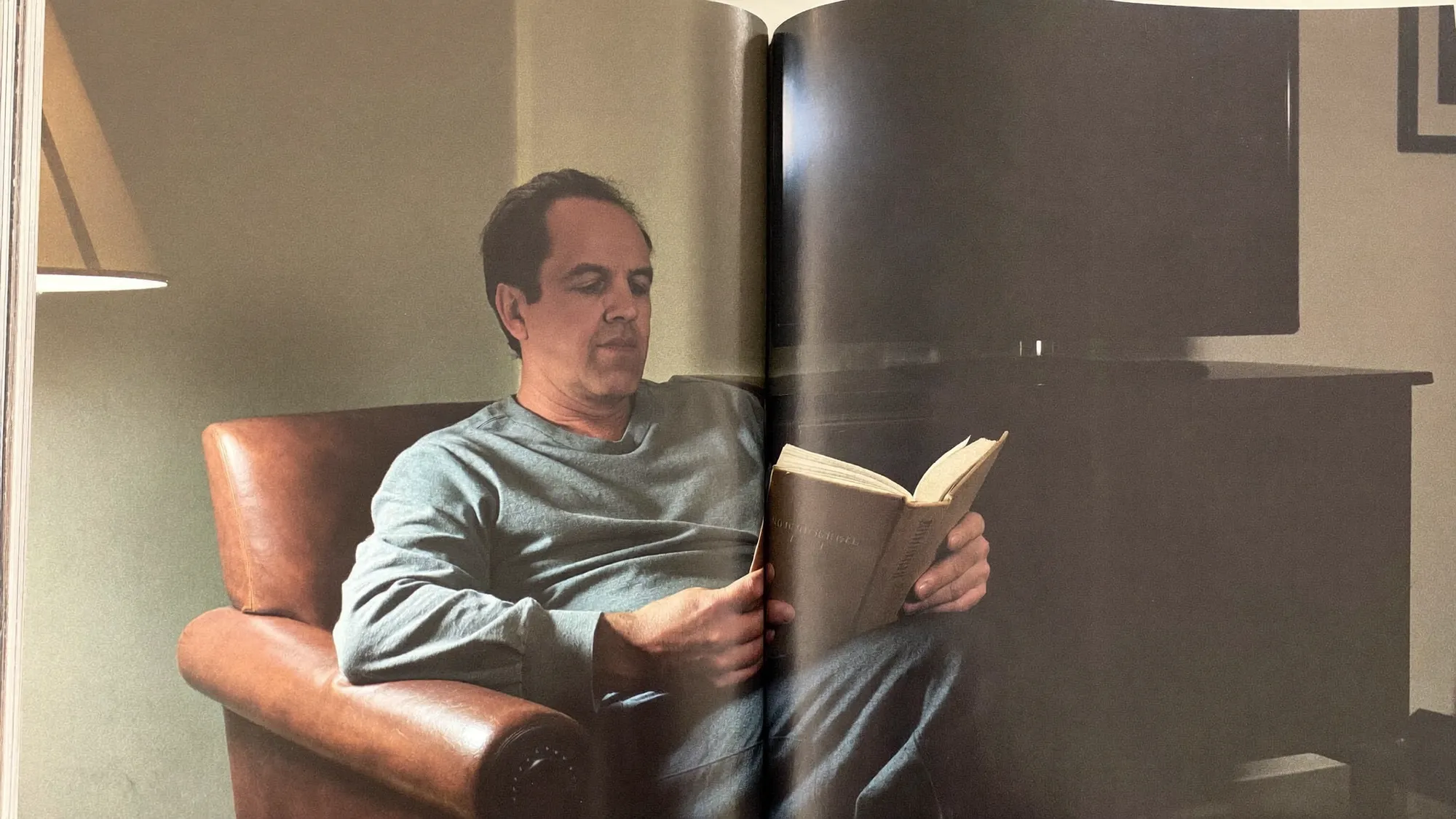 A man reading a hardcover book in an armchair by lamplight, a dark television behind him