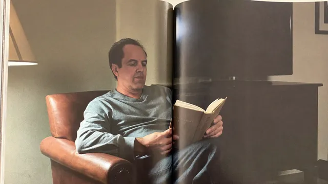 A man reading a hardcover book in an armchair by lamplight, a dark television behind him